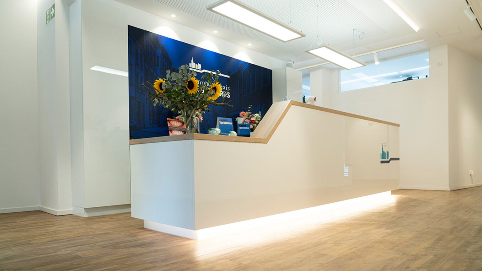 Modern gestalteter Empfangsbereich einer Zahnarztpraxis mit heller Theke, stimmungsvoller LED-Unterbeleuchtung, Holzoptik-Boden und dekorativem Blumenschmuck vor einem dunkelblauen Hintergrund mit Praxislogo.