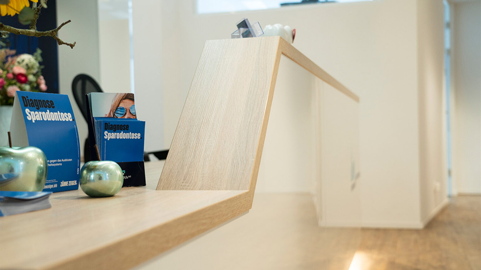 Detailaufnahme einer Empfangstheke mit abgeschrägter Holzoberfläche, Werbematerial zum Thema Zahngesundheit und dekorativen Objekten in Apfelform.