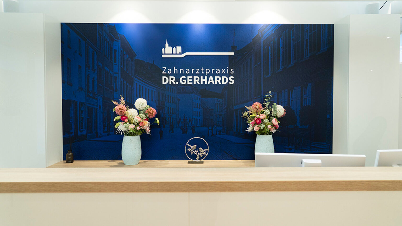 Empfangstheke der Zahnarztpraxis Dr. Gerhards mit großem blauen Wandbild, zwei Vasen mit Blumen und moderner Beschilderung.