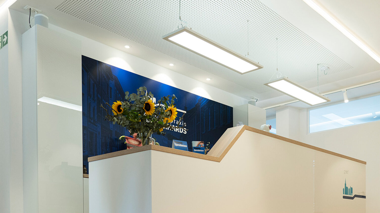 Modern gestalteter Empfangsbereich einer Zahnarztpraxis mit heller Theke, stimmungsvoller LED-Unterbeleuchtung, Holzoptik-Boden und dekorativem Blumenschmuck vor einem dunkelblauen Hintergrund mit Praxislogo.