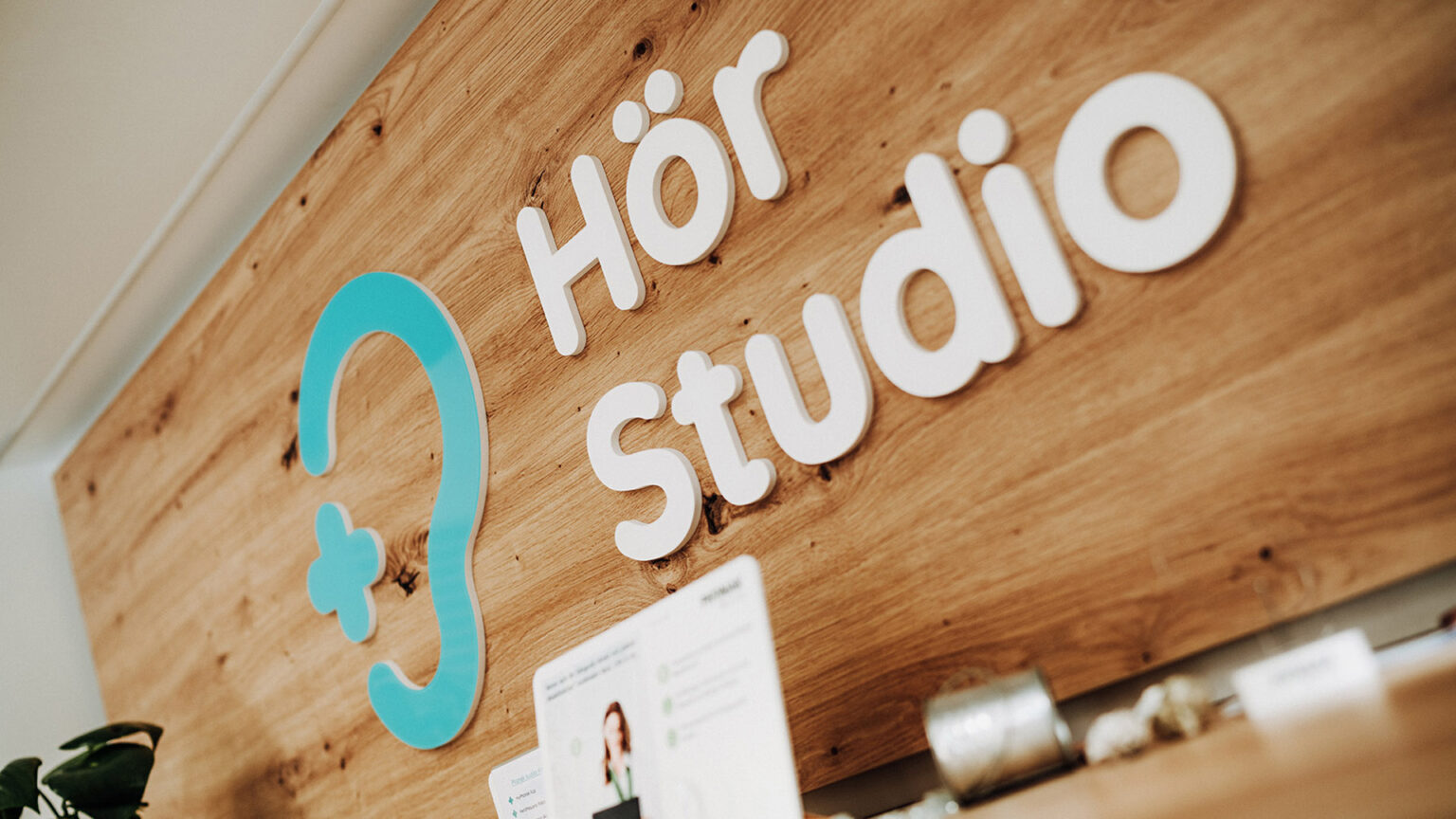 Nahaufnahme des Logos vom Hör Studio auf einer Holzrückwand. Der weiße Schriftzug „Hör Studio“ wird ergänzt durch ein stilisiertes Ohrsymbol in Türkis.